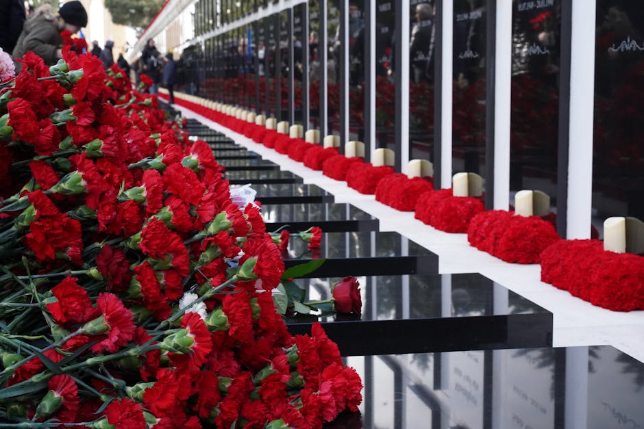 A memorial site featuring a row of white candles placed on the ground, interspersed with red carnation flowers arranged in a line along the edge of a black polished surface, possibly a memorial plaque